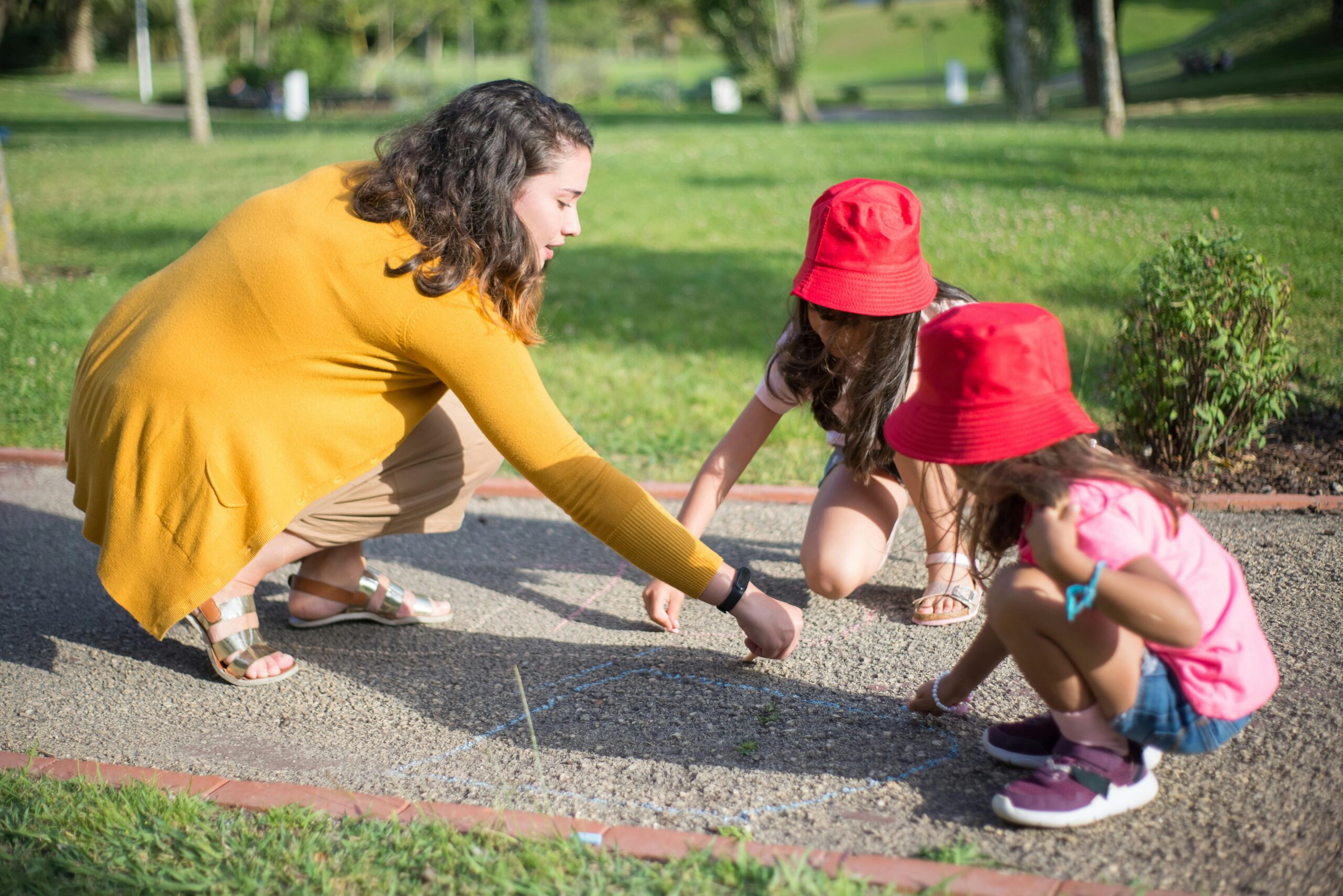 A nanny and two children drawing with chalk on a sunny day outdoors.